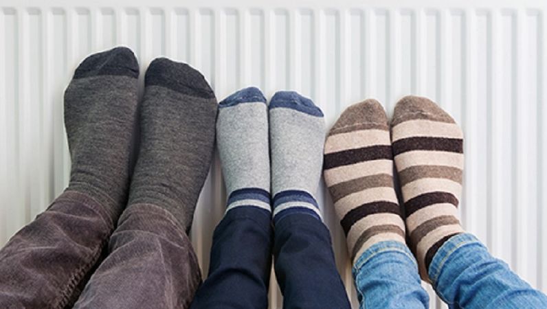 Symbolbild klimafreundlich Heizen: Zwei große Menschen und ein kleiner Mensch halten ihre Füße mit bunten Socken an einen Heizkörper, um diese zu Wärmen. 