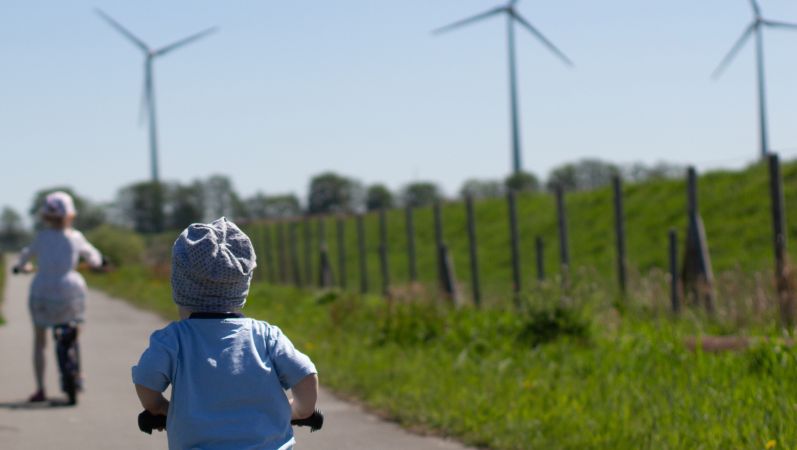 Kinder fahren mit ihren Fahrrädern auf einem Weg in Richtung Windenergieanlagen.