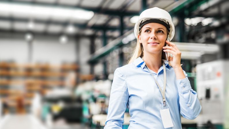 Eine Ingenieurin mit Bluse und Helm in einer Werkshalle, am Telefon.