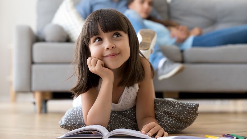 Ein Mädchen liegt mit einem Buch auf einem Holzfußboden, im Hintergrund eine Couch mit den Eltern.