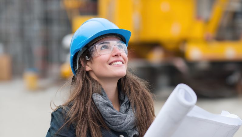 Eine Frau mit Schutzhelm hält einen Bauplan in der Hand und schaut schräg nach oben.